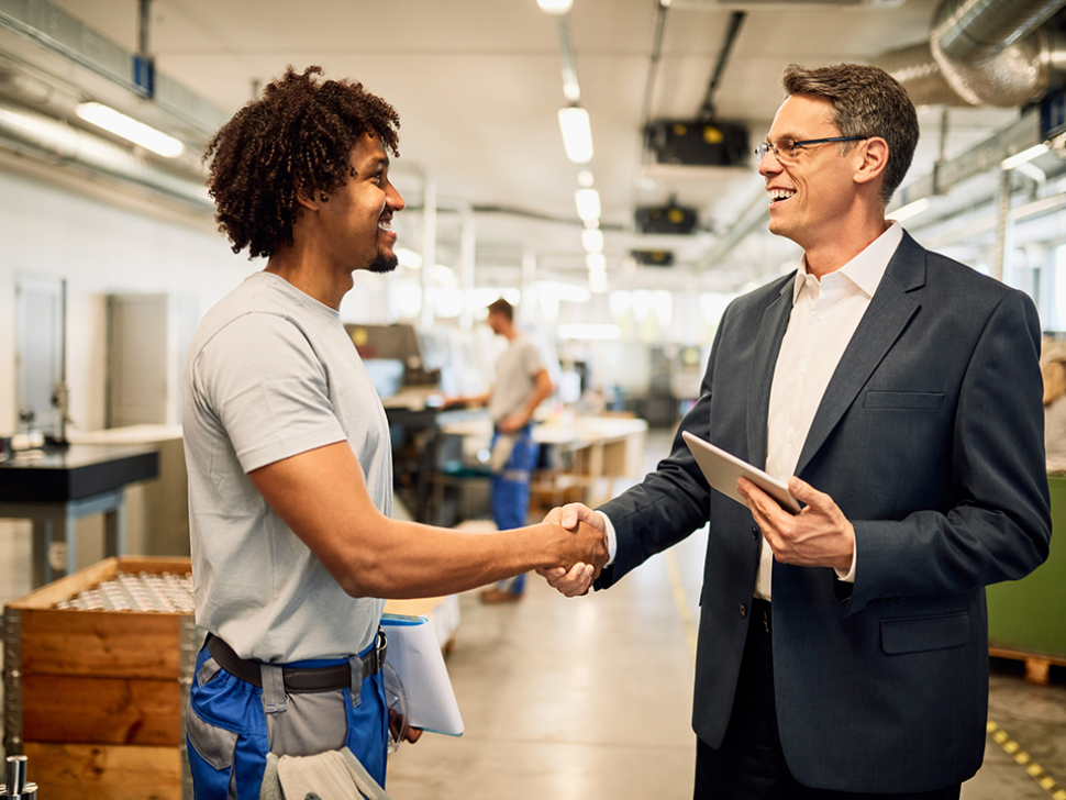 Happy engineer and young black worker handshaking while greeting in industrial building.